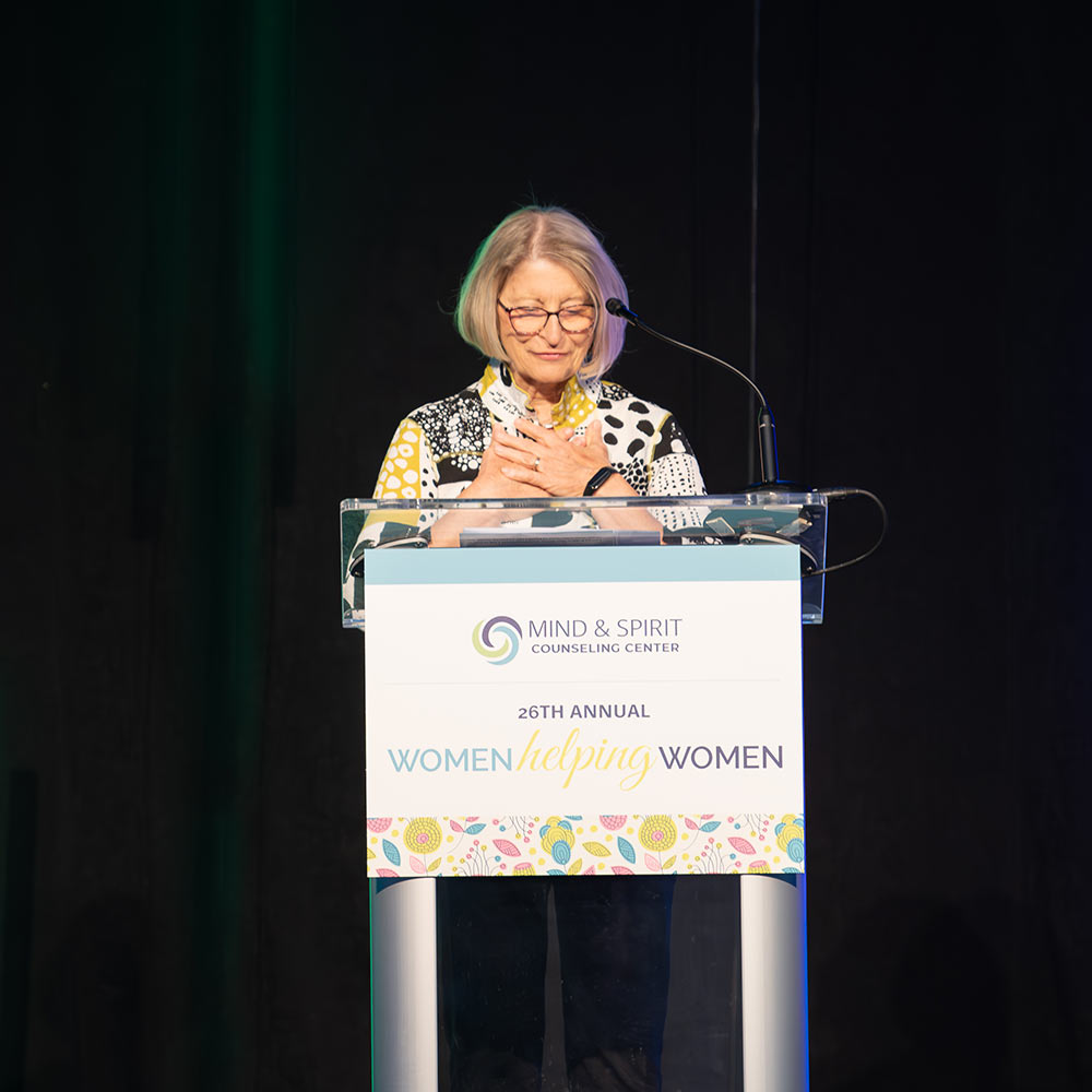 Honoree Carol Bodensteiner holds her hands to her heart while speaking at Mind & Spirit Counseling Center's 2024 Women Helping Women luncheon