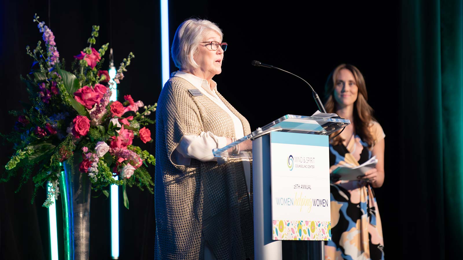 Laurie Betts Sloterdyk, retiring director of development, speaks to the crowd as Heather Stuyvesant, new director of development, stands beside her on stage at Mind & Spirit Counseling Center's 2024 Women Helping Women luncheon